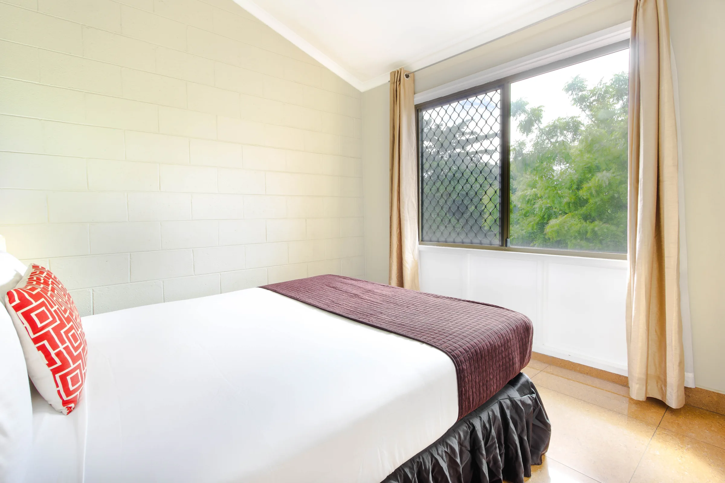Made bed beside a window with a red patterned cushion